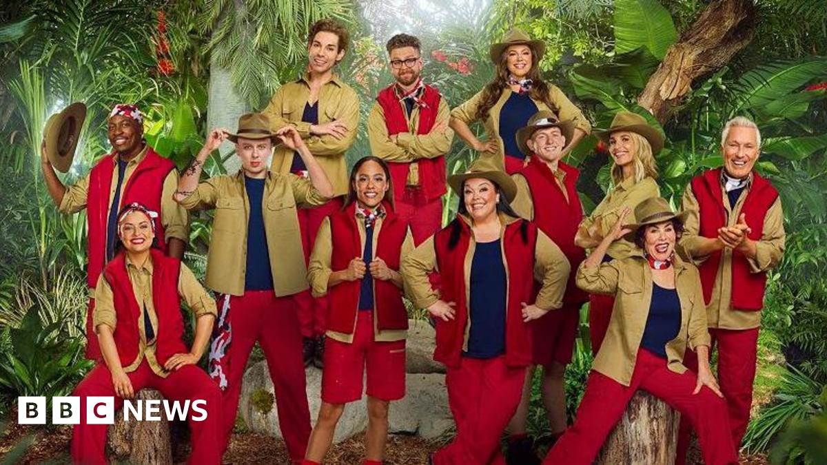 Twelve people can be seen wearing various outfits of red and khaki green, with some wearing large khaki hats. They are posed in front of a jungle backdrop as part of the announcement for the new cast of I'm a Celebrity...Get Me Out of Here!