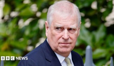 Image of Andrew Mountbatten Windsor. He is looking off to the right. Andrew has thinning grey hair and is wearing a navy suit jacket, white shirt and a yellow patterned tie