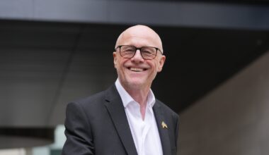 Breaking news event with a large, diverse crowd gathering in a city square under clear skies, capturing public interest.