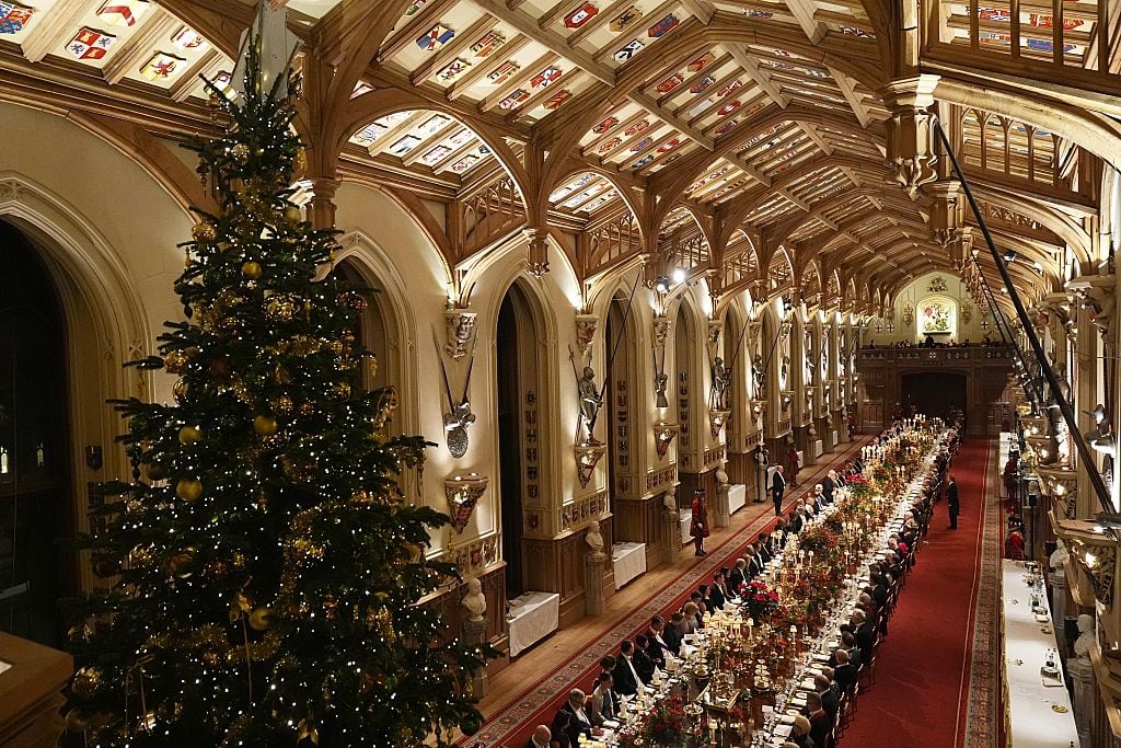 A large Christmas tree sits at the end of a large hall where dinner guests are seated at a long banquet table.