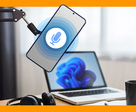 A smartphone with a microphone icon in a microphone arm, with a laptop, headphones, and a coffee mug on a desk in the background.