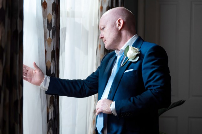 Max looks out of a window while wearing a suit in EastEnders
