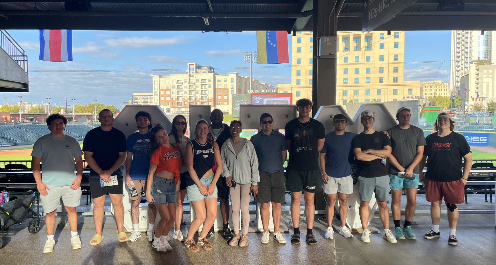 A group of people stand in a line under a covered outdoor structure at a sports venue, with a city skyline and flags visible in the background.