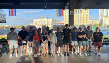 A group of people stand in a line under a covered outdoor structure at a sports venue, with a city skyline and flags visible in the background.