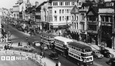 A black and white photo of Cardiff city centre. There are lines of shops on one side, castle green on the other. Through the middle is a road with double decker buses and cars on it and there are lots of people milling about.
