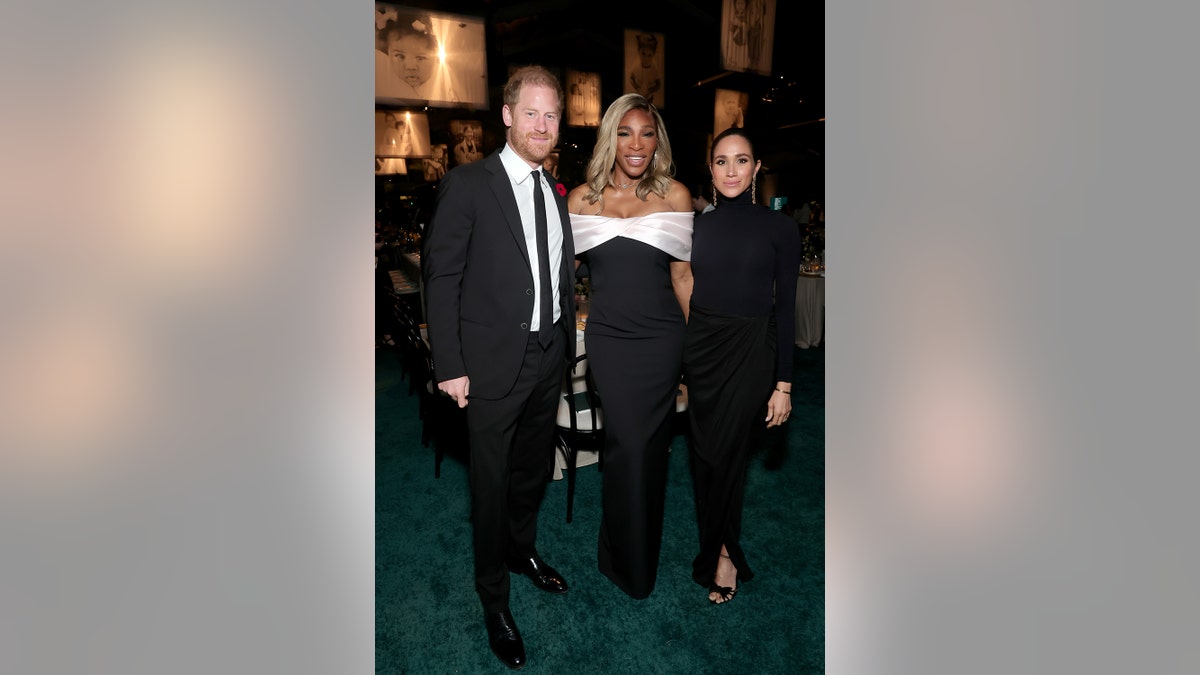 Prince Harry in a dark suit and tie posing next to Serena Williams and Meghan Markle as they wear glamorous dark gowns.