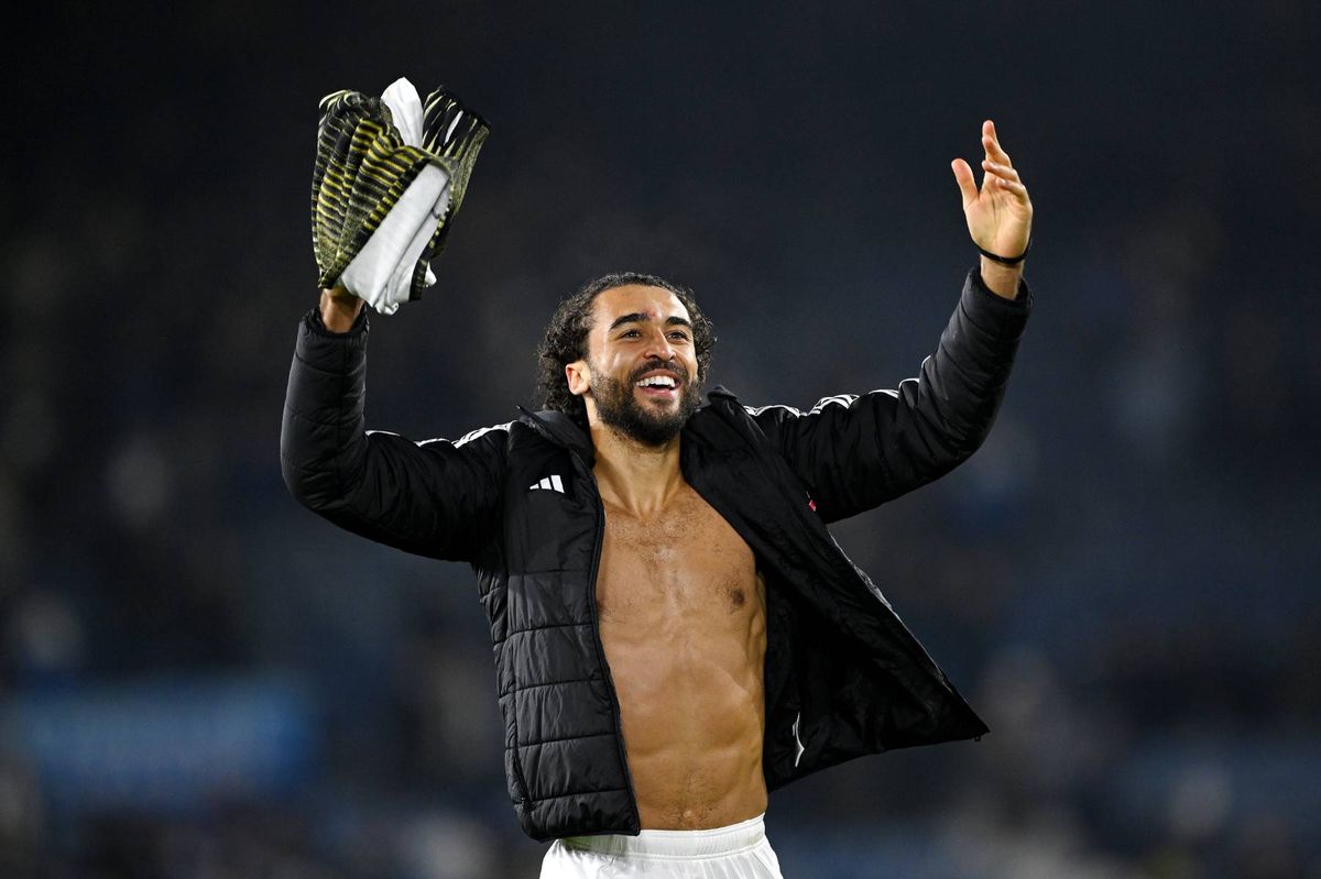 An individual, partially shirtless, raises his hands while holding a trophy, standing in an open field with a blurred background, likely during a celebratory event.