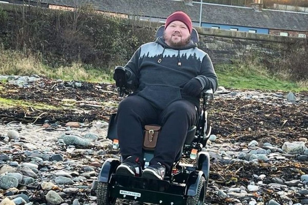 An image of a young man on a beach wearing a hoodie, trousers and trainers with a red hat, sitting in an electric wheelchair.