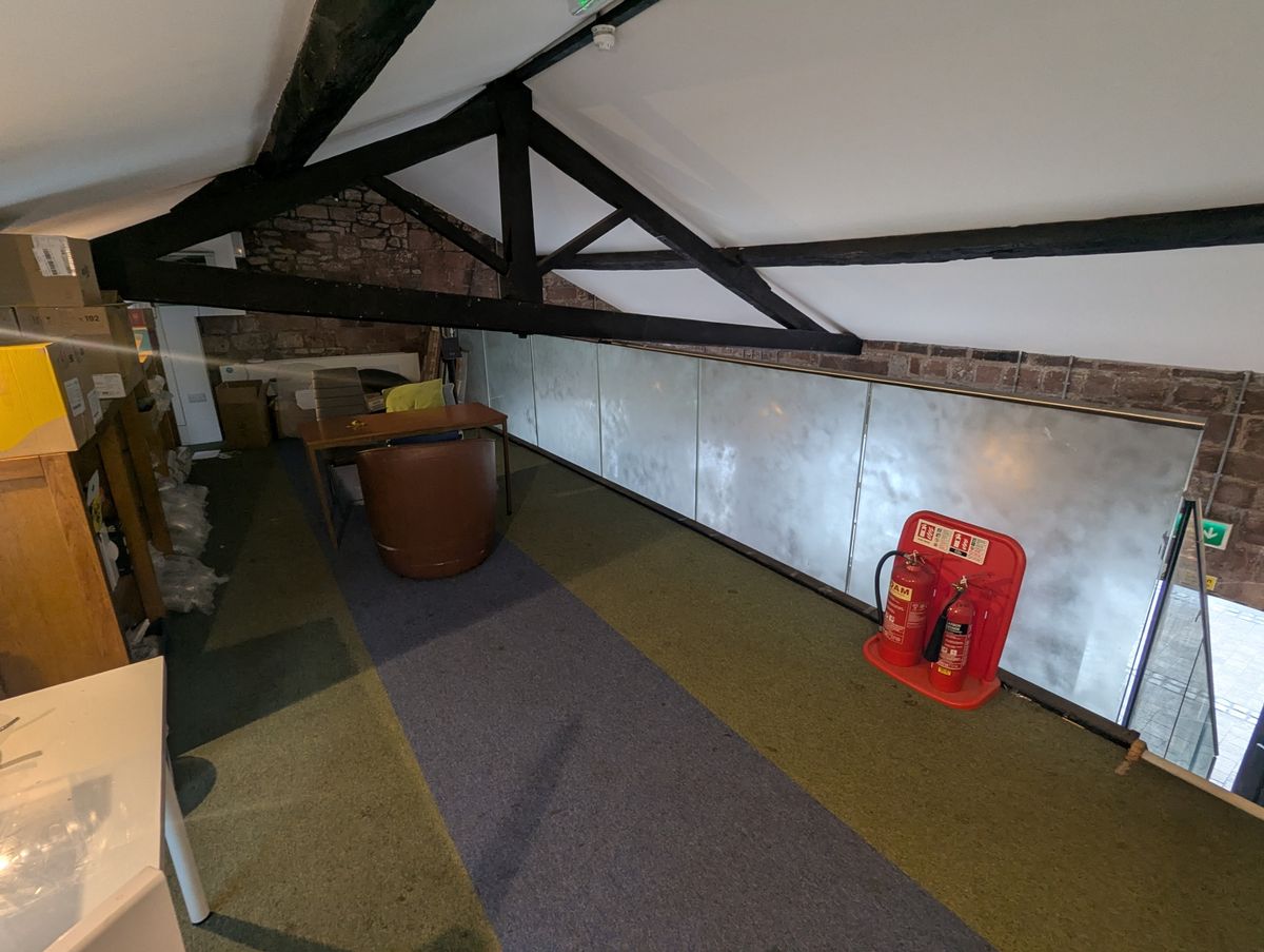 A small mezzanine space with a yellow and blue carpet. A fire extinguisher is positioned by the stairs down. A desk and several boxes are the only furnishings in the small space, which is divided by a low hanging wooden beam from the roof.