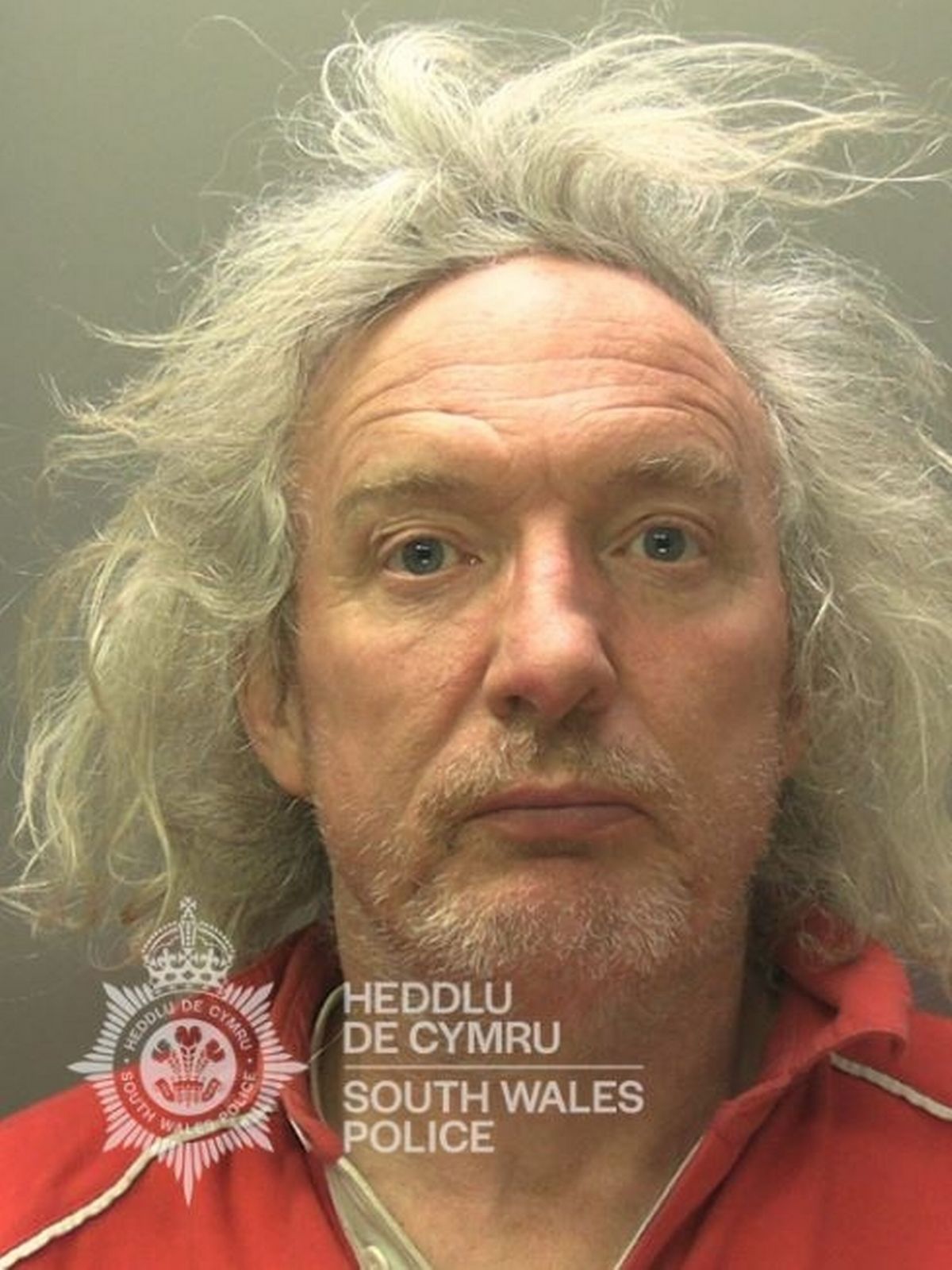 Man in his 60s with long grey hair. He is wearing a red polo shirt and has some stubble