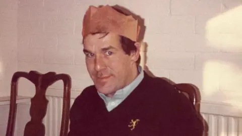 Rob Parsons Photograph of Ronnie Lockwood, who wears a red paper party hat typically worn on Christmas day. He wears a blue shirt with a navy v neck jumper over the top of it. He has bright blue eyes and brown hair and smiles at the camera. 