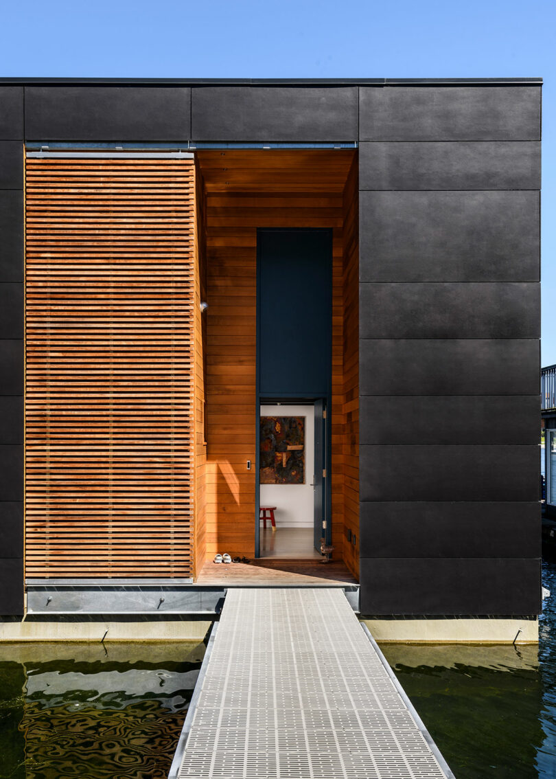 A modern floating house with dark exterior panels, wooden slats, and a metal walkway leading to the entrance over water.
