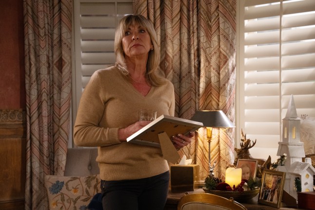 Kim stands with a glass in her hand and a book in another while at Home Farm in Emmerdale
