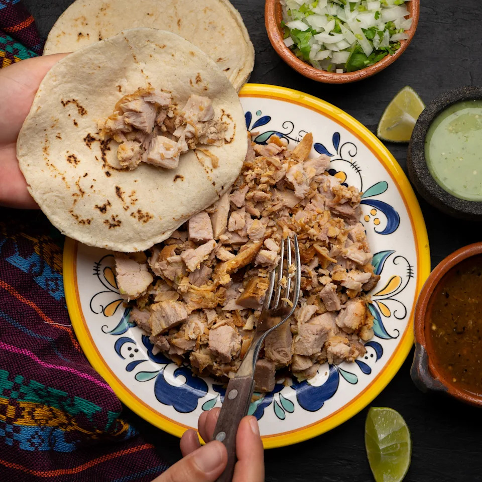 Person assembling a taco with seasoned meat, garnished with chopped onions and cilantro. Accompanied by green and red sauces, lime wedges, and tortillas