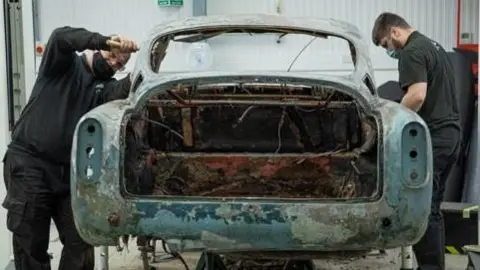 Marc Bow/Timbuktu Content Two technicians stand either side of the damaged chassis of the Aston Martin as they begin to repair it in a workshop