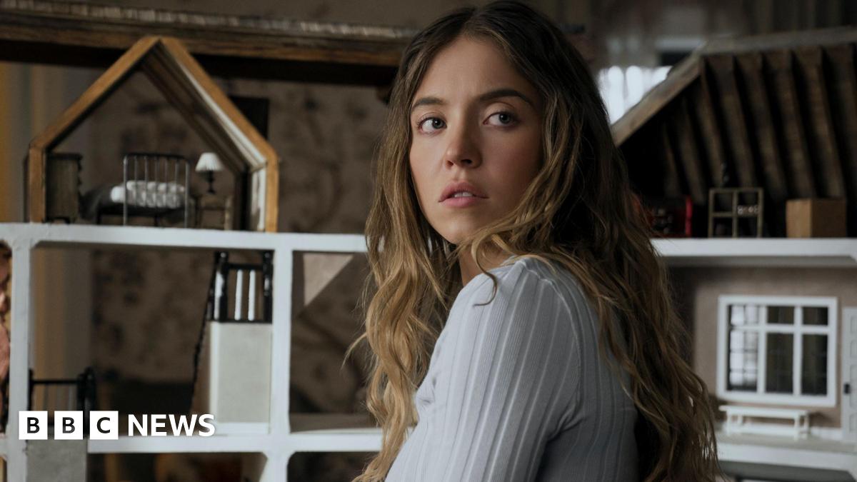 Sydney Sweeney appears with her head turned to face away from the camera. She is wearing a white long-sleeved ribbed top and is in front of a dolls house.