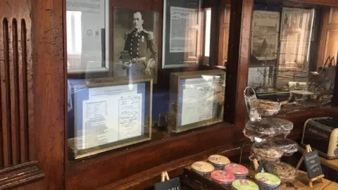 A glass cabinet with framed documents and pictures in it. In front is a table where porridge pits and cereals are laid out.
