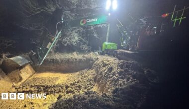 A digger removing what appears to be sand from a wooded area at night