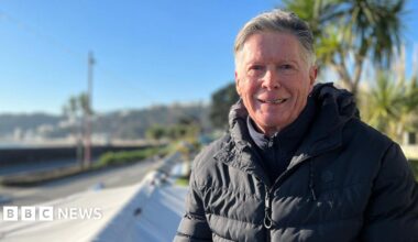 Gordon is wearing a black puffer jacket sits outdoors on a folded sail or tarp near a wooden fence, with palm trees and a clear blue sky in the background. The setting appears coastal, with water and distant hills visible.