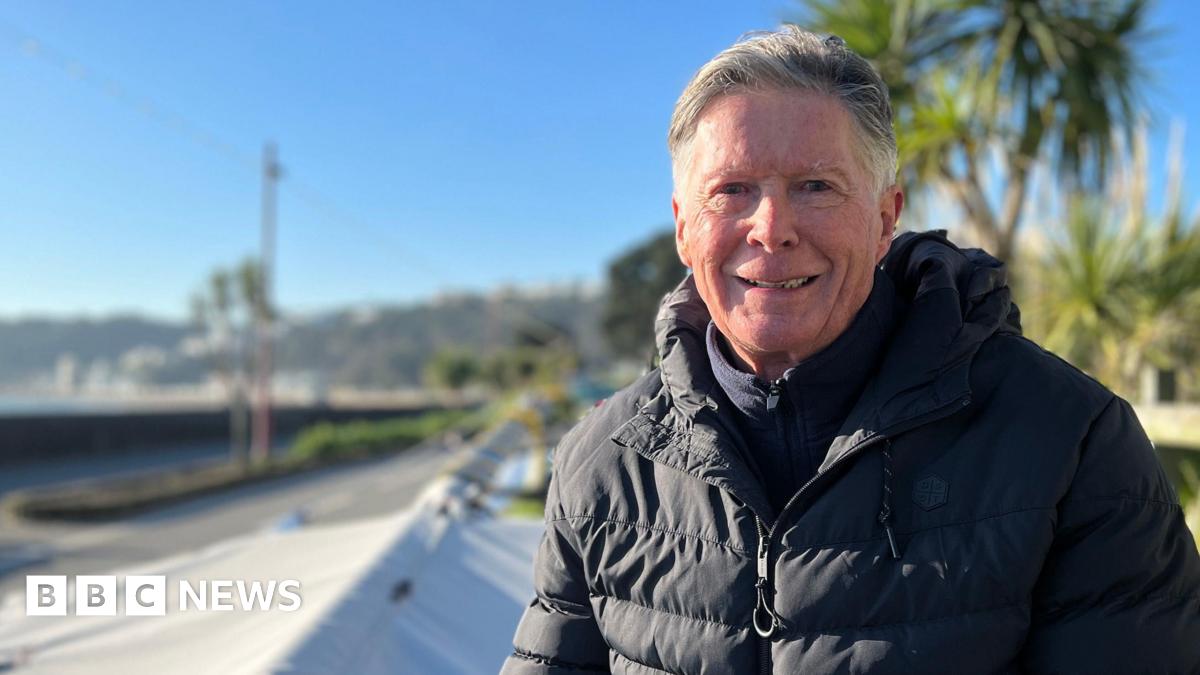 Gordon is wearing a black puffer jacket sits outdoors on a folded sail or tarp near a wooden fence, with palm trees and a clear blue sky in the background. The setting appears coastal, with water and distant hills visible.