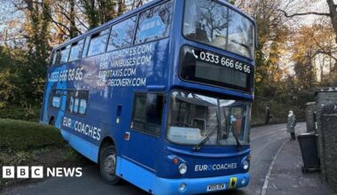 A blue double decker bus with a number along the top and the words  "Eurocoaches, Eurominibuses, Eurotaxis" on the side. Its front end is blocking a narrow road, which bends to the right behind it. The back of the bus appears to have reversed over a wall/hedge off to the side of the road and is tilted upwards.