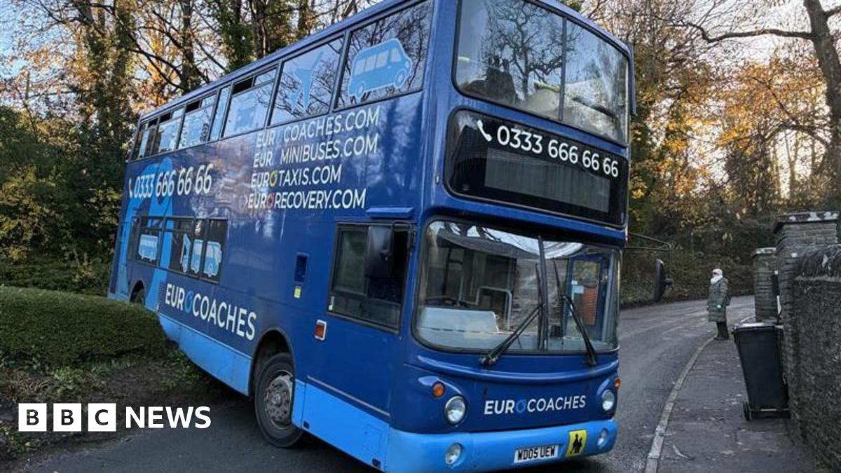 A blue double decker bus with a number along the top and the words  "Eurocoaches, Eurominibuses, Eurotaxis" on the side. Its front end is blocking a narrow road, which bends to the right behind it. The back of the bus appears to have reversed over a wall/hedge off to the side of the road and is tilted upwards.
