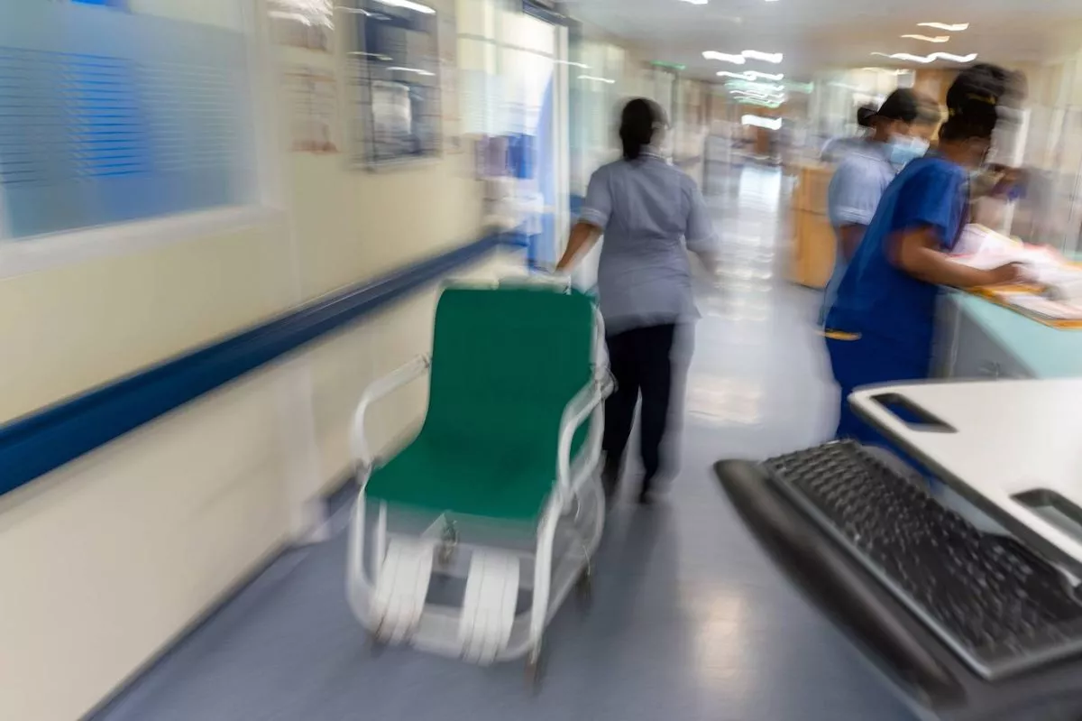 In a hospital corridor, several medical staff members are seen moving about. One individual is pushing a medical cart equipped with a wheelchair, while others are engaged in various tasks. The corridor is well-lit, with walls painted in light colors, and there are medical devices and equipment visible in the background.
