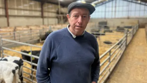 A farmer in a navy jumper is standing in front of a number of cows in a large shed. He's also wearing a cap.