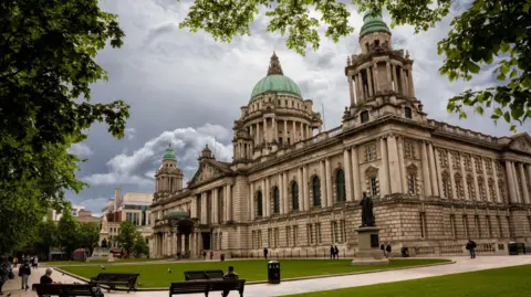 Getty Images The exterior of Belfast City Hall. It is a large, white Victorian building with blue-domed roofs. There is a large statue of Queen Victoria outside of the main entrance. There is a large statue of an old man to the right of the building, which sits on a neat square of green grass. 
