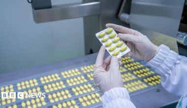 Gloved hands hold a tray of yellow tablets, above rows of yellow tablets on a conveyor belt.