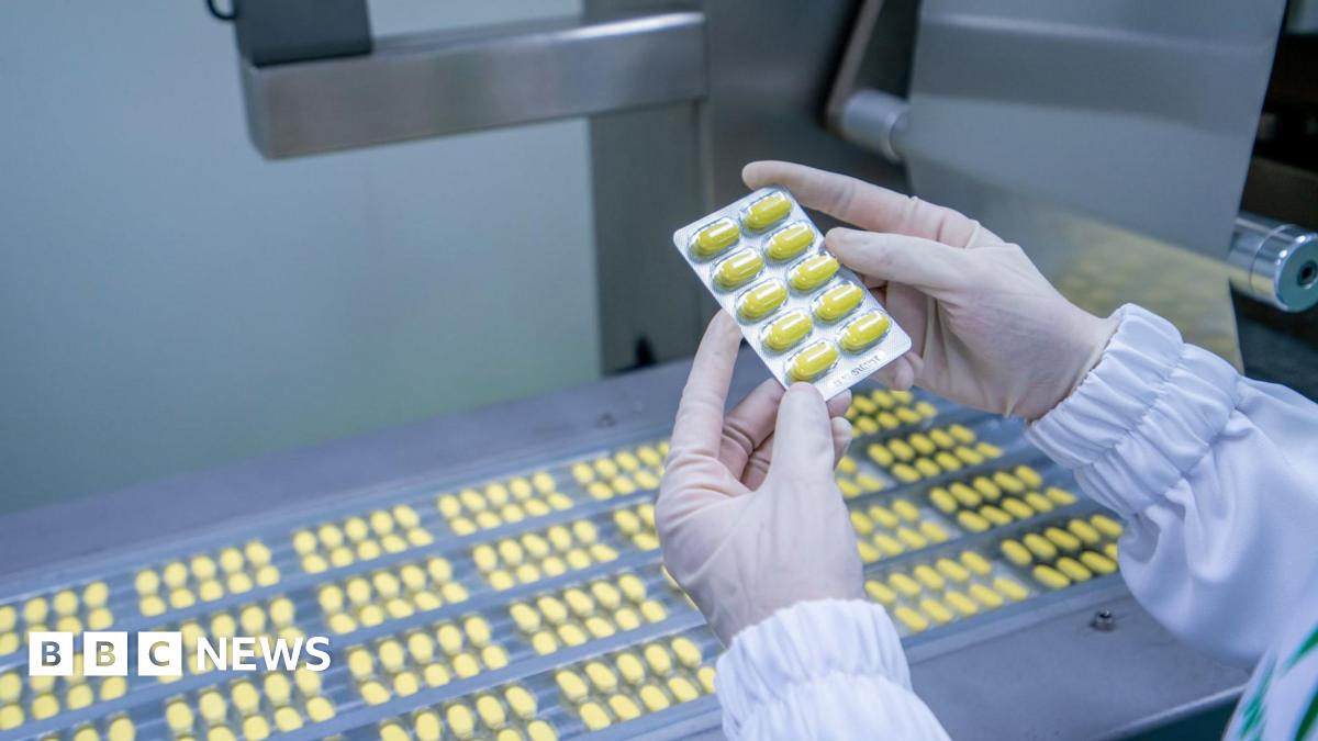 Gloved hands hold a tray of yellow tablets, above rows of yellow tablets on a conveyor belt.