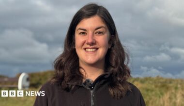 Niamh McDevitt pictured with shoulder length dark brown hair smiling at the camera with teeth. She has a septum piercing. She is wearing a black zip up fleece with the Alderney Wildlife Trust logo on.