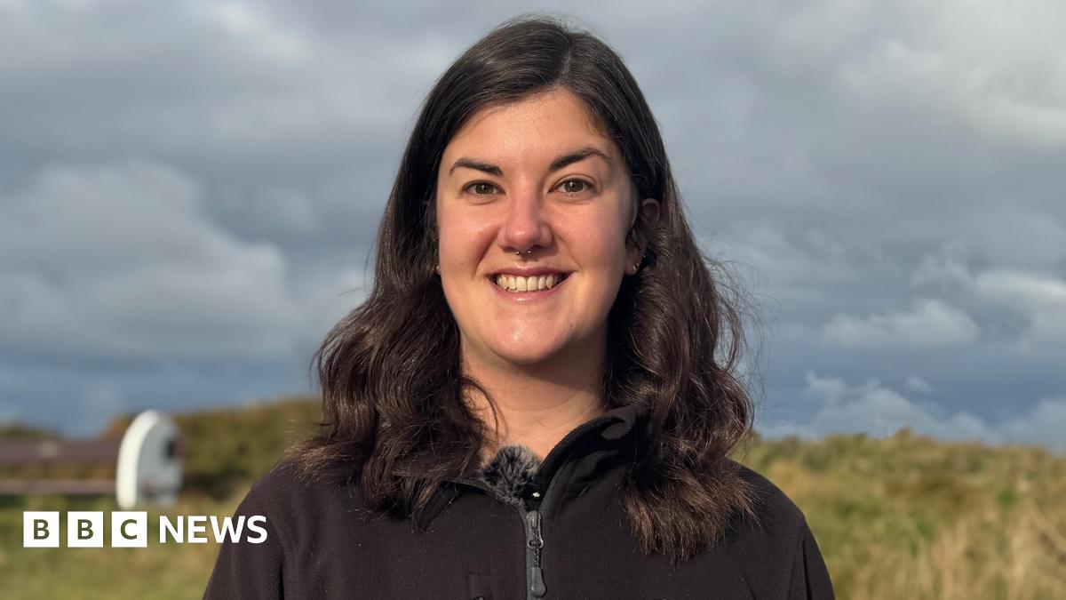 Niamh McDevitt pictured with shoulder length dark brown hair smiling at the camera with teeth. She has a septum piercing. She is wearing a black zip up fleece with the Alderney Wildlife Trust logo on.