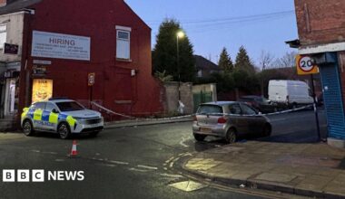 A street at dusk showing a police car parked on the left side near a red brick building with a large white sign. The area is cordoned off with police tape, and traffic cones are placed on the road. A silver car is parked on the right side of the street, and a white van is visible further down the road. There are 20 mph speed limit signs on both sides of the street.