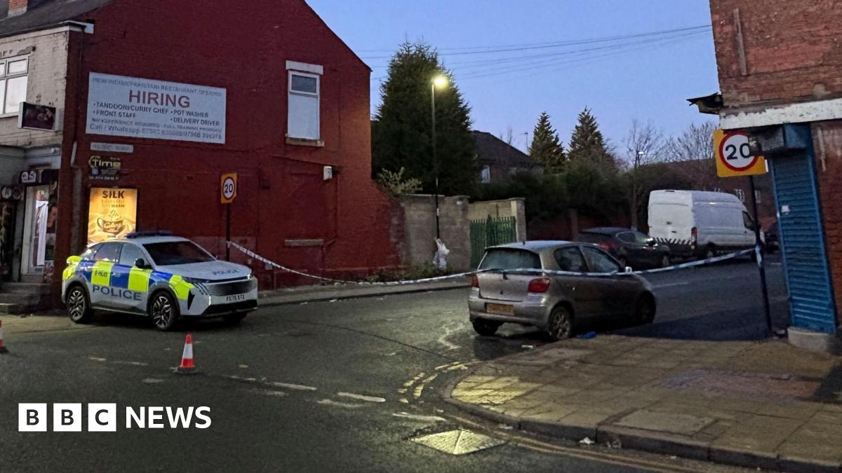 A street at dusk showing a police car parked on the left side near a red brick building with a large white sign. The area is cordoned off with police tape, and traffic cones are placed on the road. A silver car is parked on the right side of the street, and a white van is visible further down the road. There are 20 mph speed limit signs on both sides of the street.