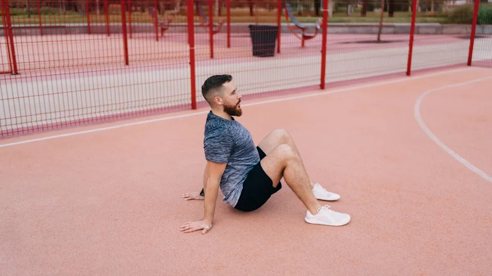 Man in a park sitting down with knees bent and hands placed behind his hips.