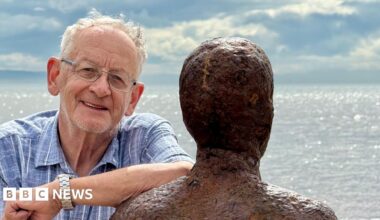 Ron Davies with grey hair wearing a blue and white shirt and glasses leans on one of the Iron Men sculptures on Crosby beach with the incoming tide in the background on a clear day. He is smiling.