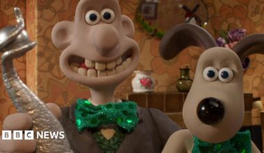 Image of clay modelled characters Wallace and his dog Gromit.  Wallace is holding a silver trophy.  They are both wearing green shiny green bow ties.  In the background can be seen a tile fireplace, oval mirror above the fire and a china and glass vase.