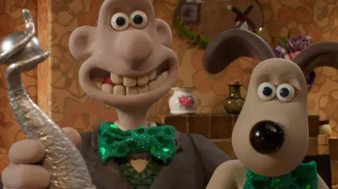 PA Image of clay modelled characters Wallace and his dog Gromit.  Wallace is holding a silver trophy.  They are both wearing green shiny green bow ties.  In the background can be seen a tile fireplace, oval mirror above the fire and a china and glass vase.
