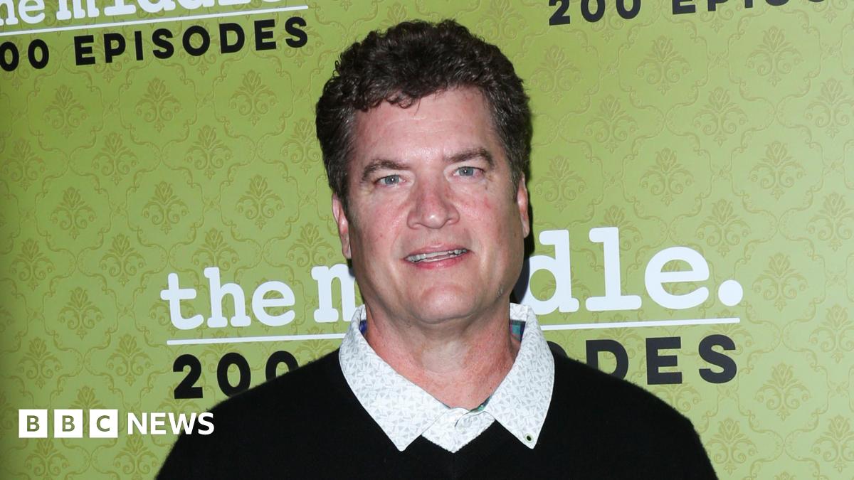 Mid shot of Pat Finn wearing a dark sweater and button-up white shirt in front of a green wallpaper backdrop ahead of a viewing of the Middle.