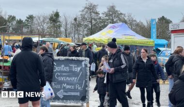 A sign says Santas Toy giveaway and free mince pie.  Stalls can be seen in the background and people are milling around wrapped up in winter coats.