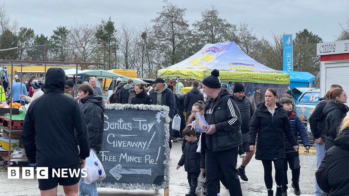 A sign says Santas Toy giveaway and free mince pie.  Stalls can be seen in the background and people are milling around wrapped up in winter coats.