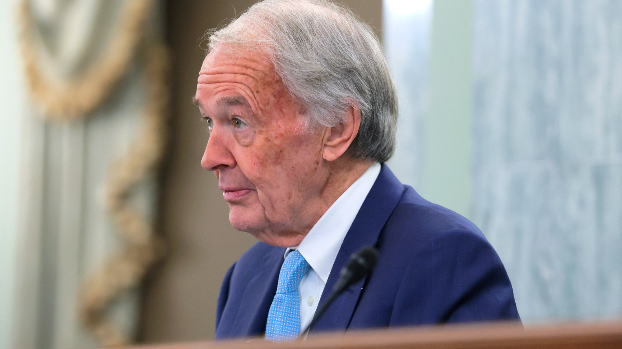 side view from the chest up of an old man wearing a dark blue suit inside a fancy room