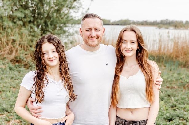 Jody, with fair skin and brown hair, has his arms wrapped around his young daughters in a rural setting. They're smiling