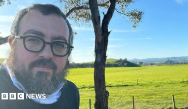 Two women comfort each other as they stare at flowers left in tribute to the victims of Sunday's shooting attack at Bondi beach. One of the women, dressed in a yellow shirt, is sitting on the road, while the other kneels next to her with her hand on her shoulder.