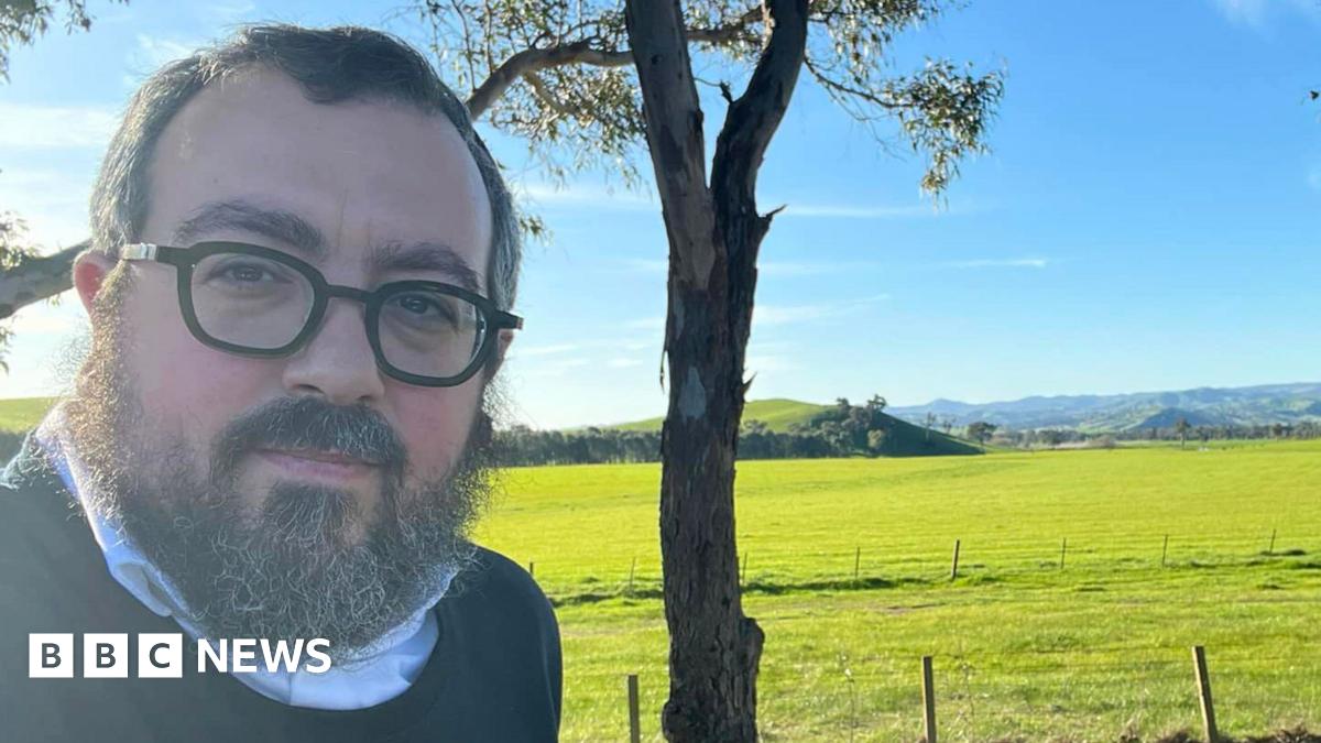 Two women comfort each other as they stare at flowers left in tribute to the victims of Sunday's shooting attack at Bondi beach. One of the women, dressed in a yellow shirt, is sitting on the road, while the other kneels next to her with her hand on her shoulder.