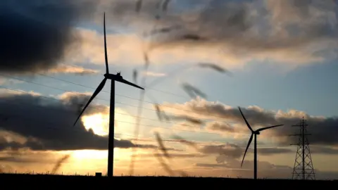 General views of a wind farm in the Scottish Highlands - two wind turbines stand in silhouette against a cloudy sky.