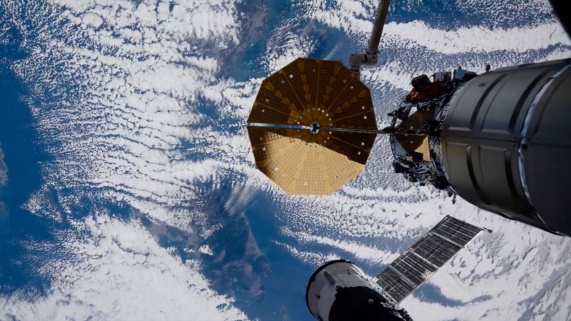 land is seen peaking through the cloud cover over a mostly blue Earth as a berthed spaceship extends into the frame from a space station