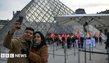 A couple take a selfie against the backdrop of strikers with placards at the Louvre Musuem