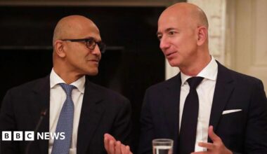 Microsoft CEO Satya Nadella (left) and Amazon founder Jeff Bezos (right), both wearing black suits over white shirts and sporting neck ties, having a conversation at an event in the US